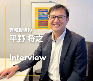 笑顔でインタビューに応じる専務取締役・平野将之氏がオフィスで座っている様子
